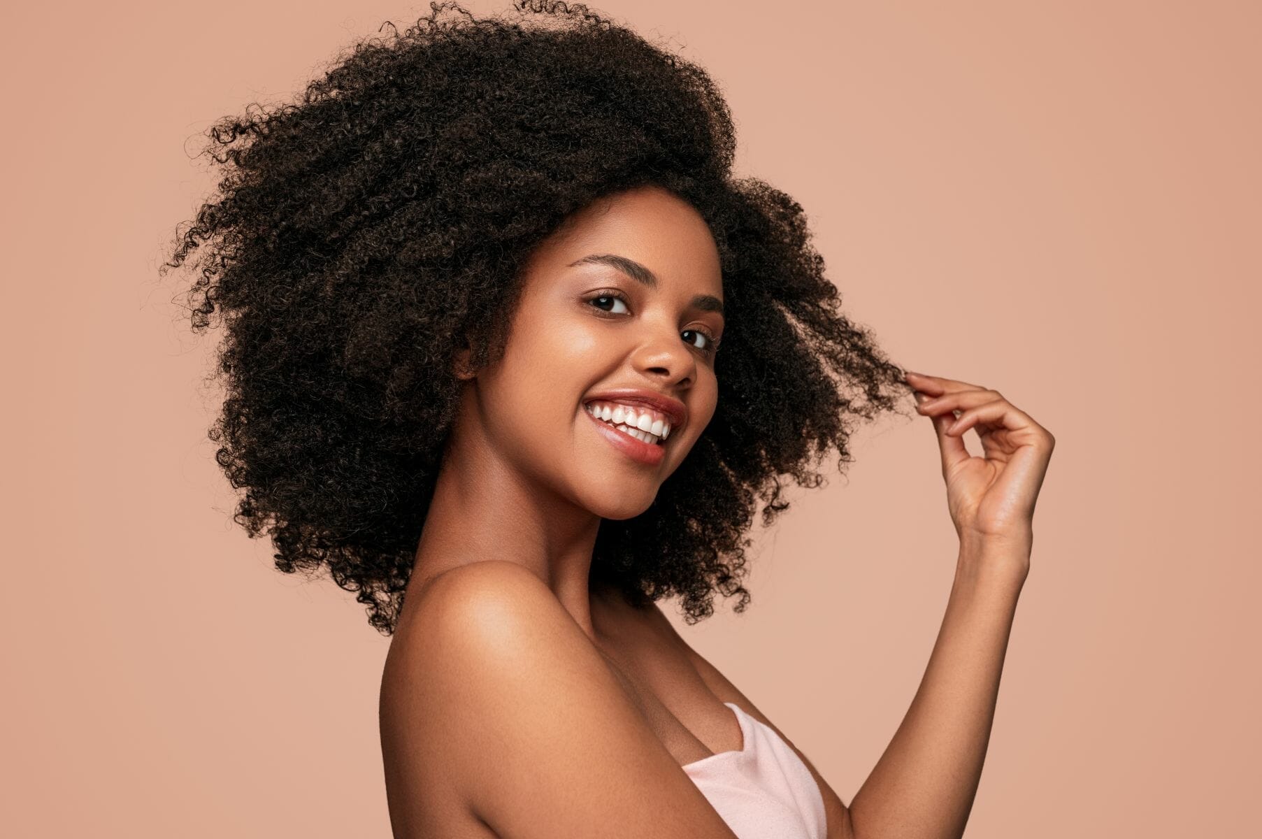 Side view of positive African American female model smiling for camera and touching clean curly hair after hygienic routine against brown background