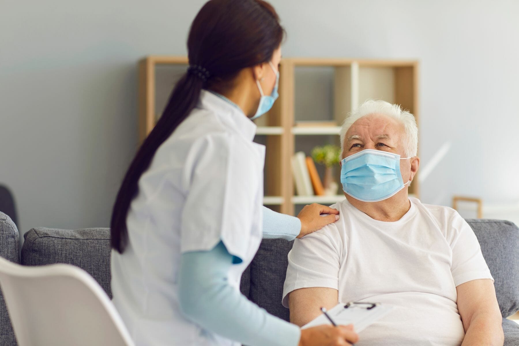 Caring female doctor supporting and cheering up senior male patient during home visit. Therapeutist and aged white-haired man, both wearing medical face masks, communicating and discussing treatment